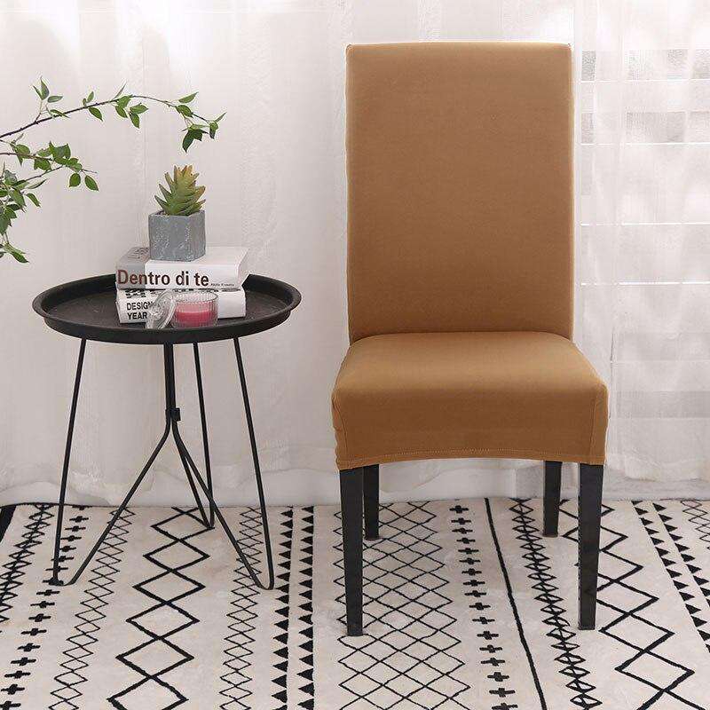 Funda de silla color camel en tela el?stica y ajustable, presentada en un entorno moderno con mesa redonda negra, planta decorativa, libros y una alfombra geom??trica, ideal para renovar el estilo de cualquier silla. Disponible en blueemoonco.com.